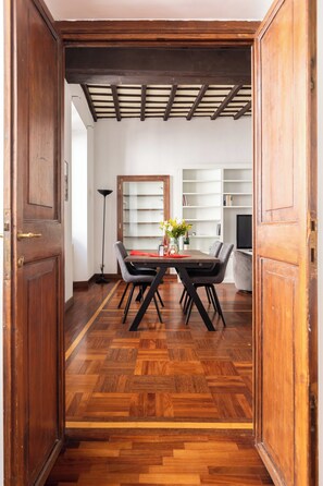 Un comedor con una mesa de madera, sillas y un florero. Se observa una estantería y un televisor al fondo.