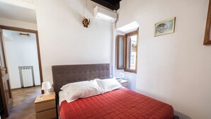 A bedroom with a red quilted bedspread, a wooden nightstand with a lamp, a bedside table, and a window with a view. - Campo De Fiori Large Family Apartment (Rome)