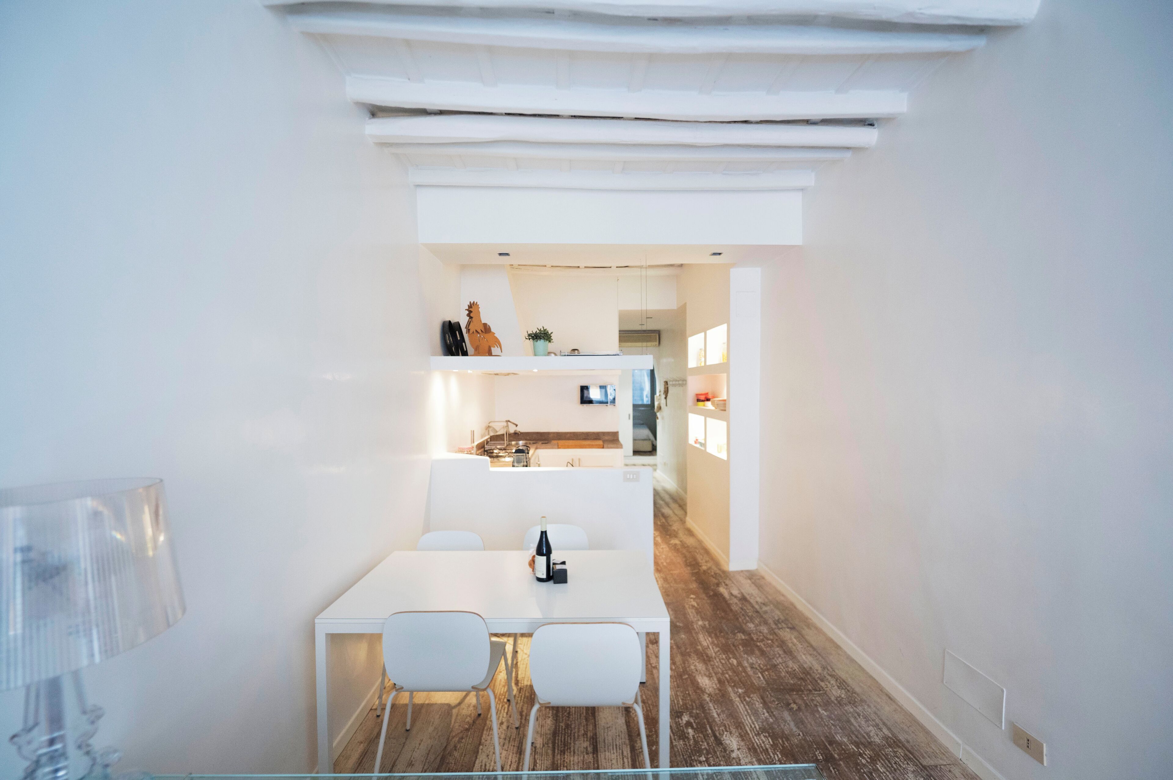 A modern, minimalist dining area with a white table and chairs, a clear glass partition, and a wooden floor.
