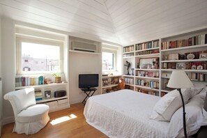 Ein Schlafzimmer mit Bett, Sessel, Fernseher, Bücherregal und Fenster.
