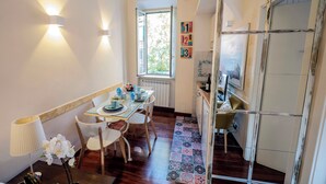 A dining area with a table set for four, a window with a view of greenery, and a kitchen area with a tiled backsplash. - Colosseum 2Br Elegance And Charm  (Rome)