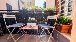 An outdoor seating area with two white folding chairs and a small table, surrounded by potted plants and a stone wall. - Colosseum 2Br Elegance And Charm  (Rome)