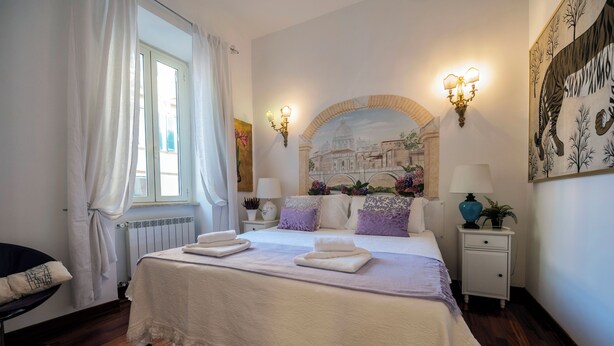 A neatly arranged bedroom with a bed, bedside tables, a chair, and a window with curtains. - Colosseum 2Br Elegance And Charm  (Rome)