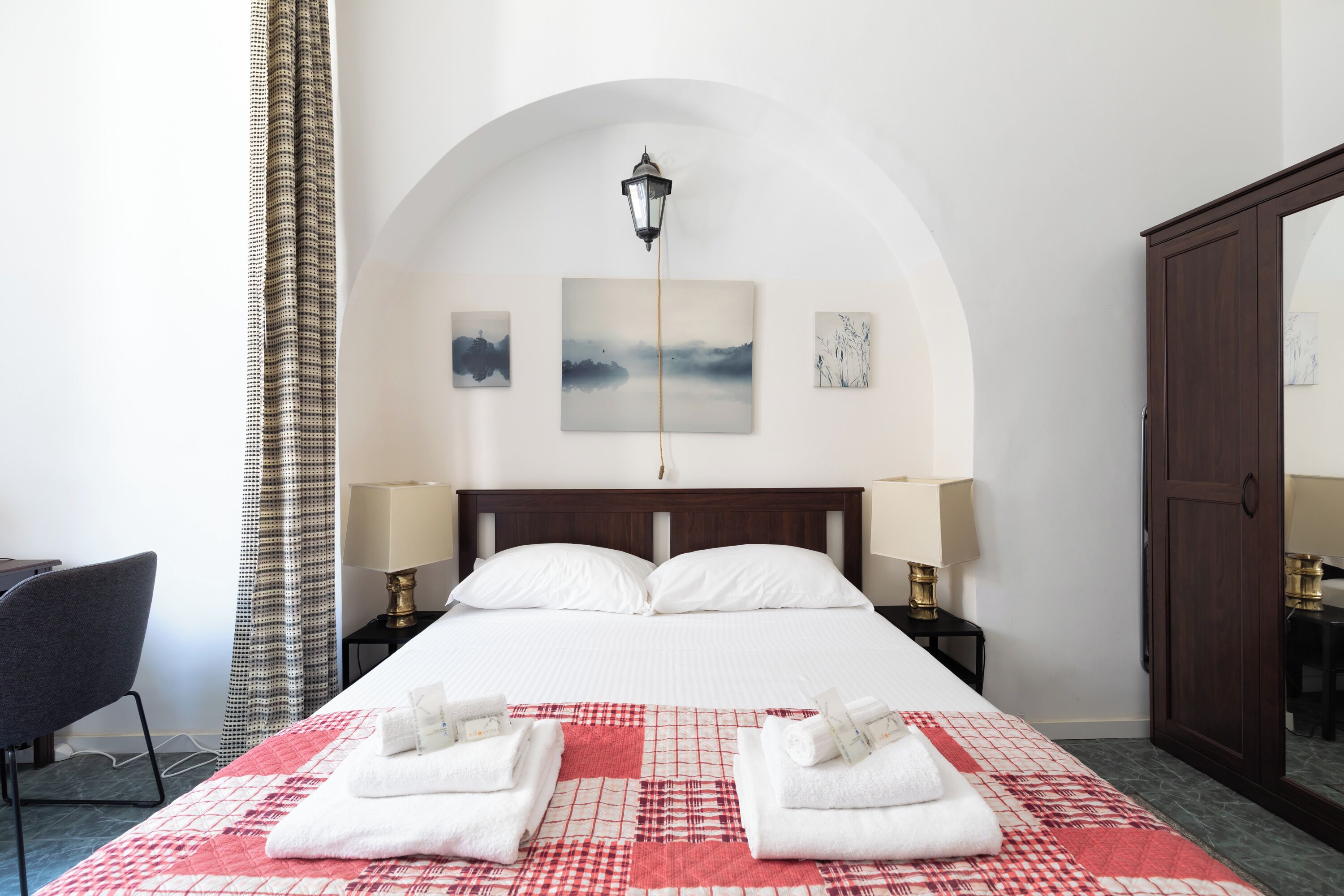A hotel room with a large bed, two bedside lamps, a nightstand, a chair, a wardrobe, and a view of the sky through an arched window.