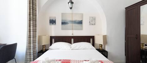 A hotel room with a large bed, two bedside lamps, a nightstand, a chair, a wardrobe, and a view of the sky through an arched window.
