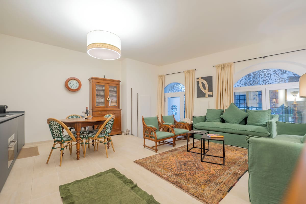 A living room with a green sofa, wooden dining table, and chairs. There is a patterned rug on the floor.