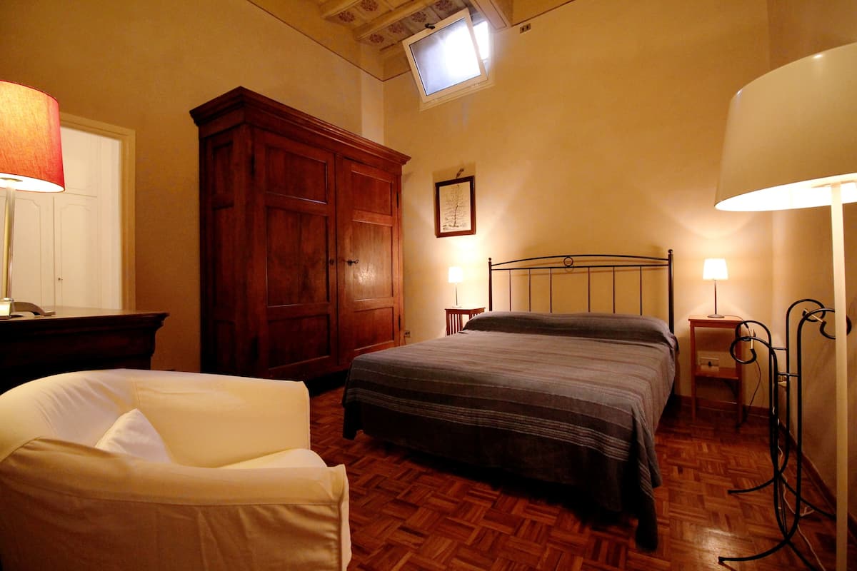 A bedroom with a bed, a wooden wardrobe, a desk with a lamp, and a chair.