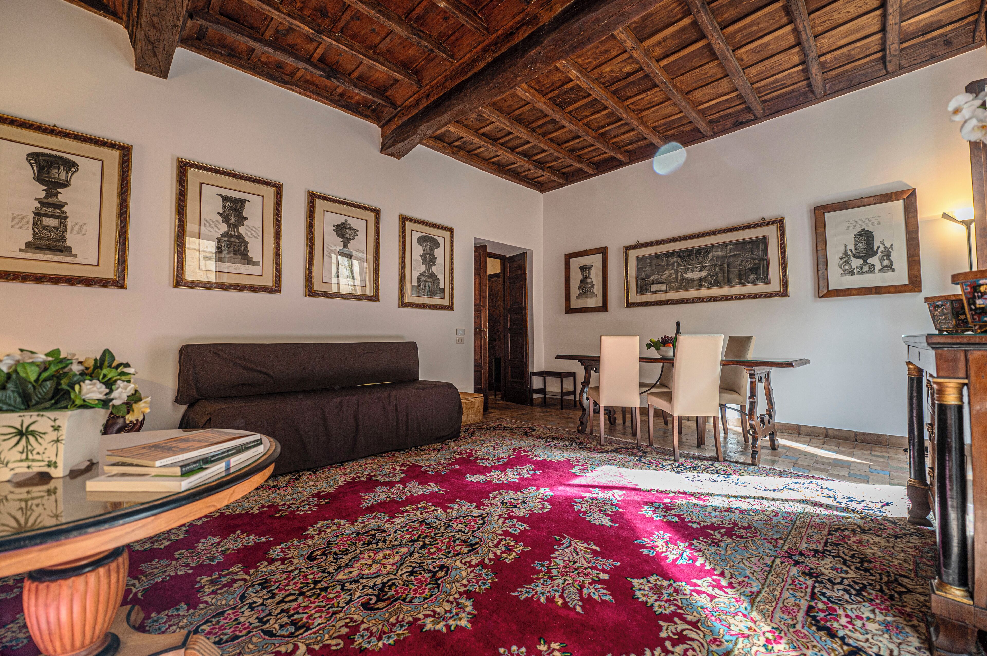 A room with a wooden ceiling, a red patterned carpet, a sofa, a dining table with chairs, and framed pictures on the walls.