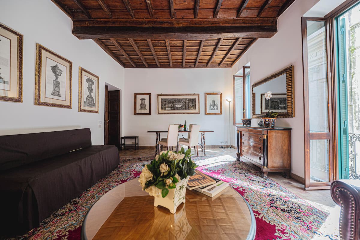 A living room with a dark-colored sofa, a wooden coffee table with a floral centerpiece, a dining area with a table and chairs, and a large mirror with a decorative frame.