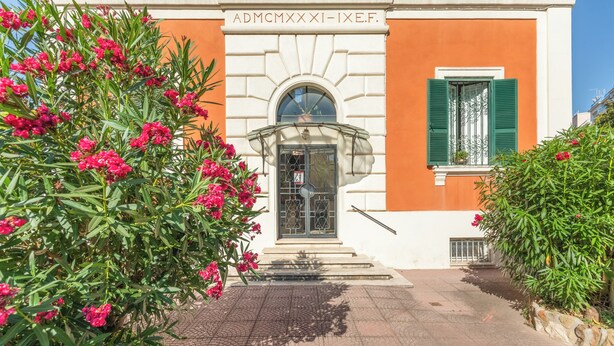 Un bâtiment à la façade blanche et aux ornements orange, doté d’une entrée avec une porte vitrée et des volets verts, le tout entouré d’une végétation luxuriante composée de plantes à fleurs roses.