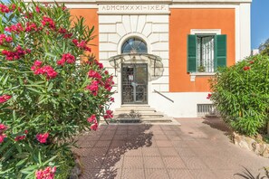Un bâtiment à la façade blanche et aux ornements orange, doté d’une entrée avec une porte vitrée et des volets verts, le tout entouré d’une végétation luxuriante composée de plantes à fleurs roses.