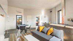 Un salon moderne avec un canapé gris, des coussins jaunes et gris, une table basse blanche et un tapis à motifs. On y trouve également un coin repas avec une table ronde et des chaises, ainsi qu’une cuisine avec un plan de travail et un évier en acier inoxydable.