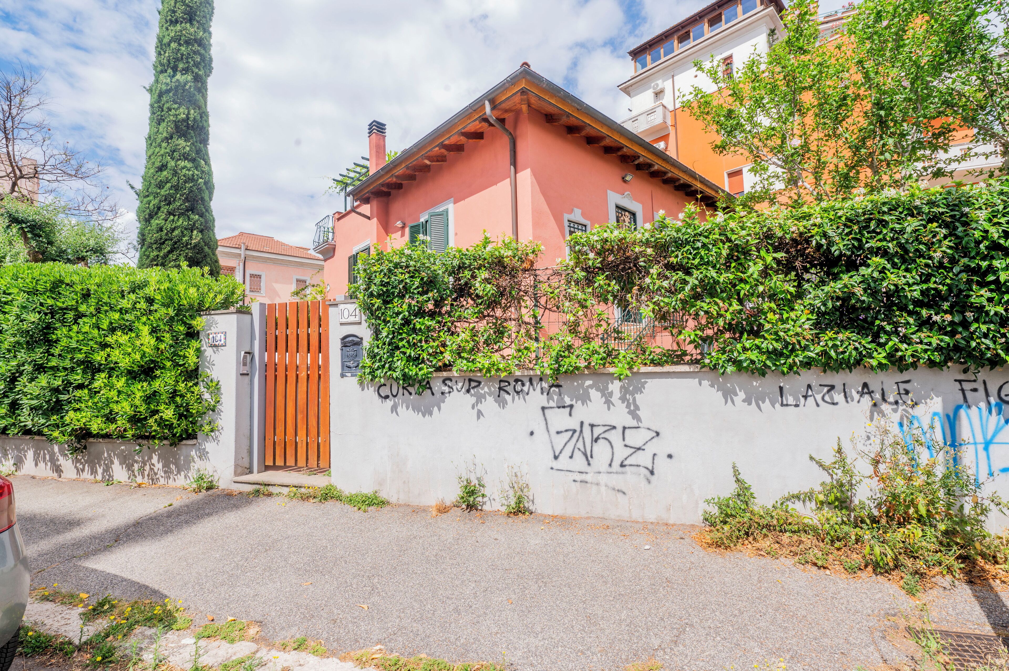 Una casa rosa con un portón de madera, rodeada de follaje y un muro con grafitis.