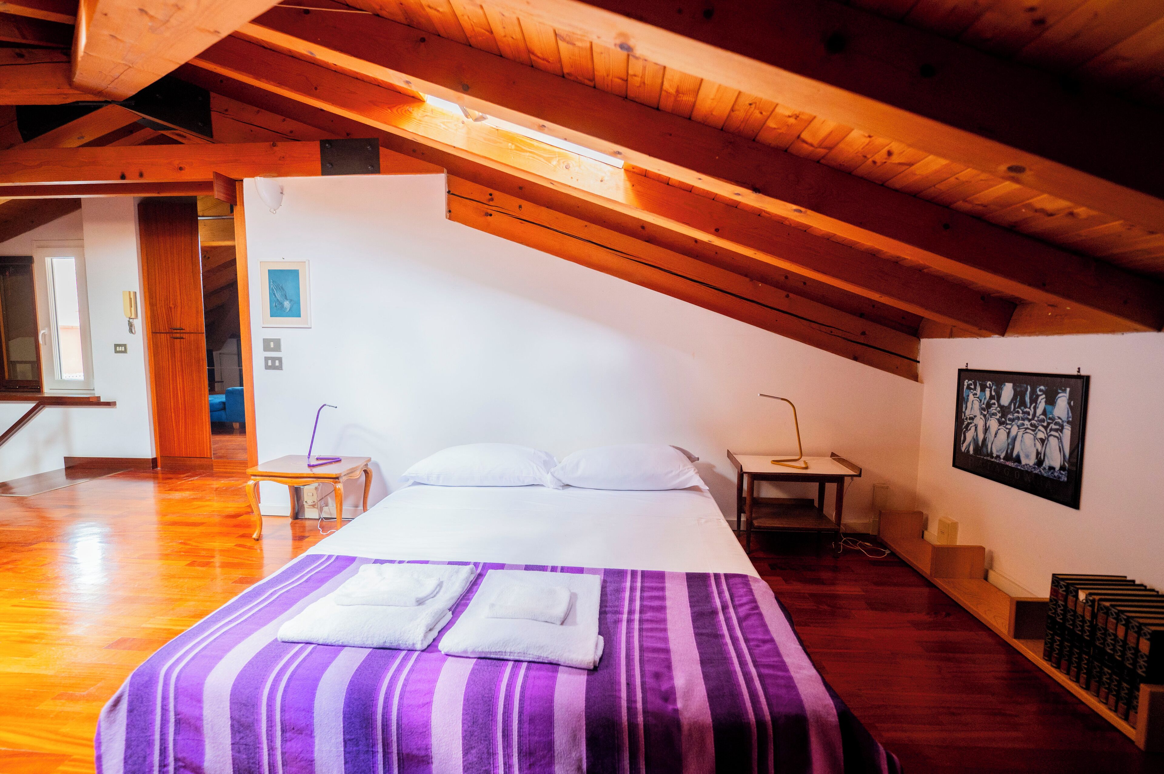 A room with a bed, wooden beams, a white wall, a picture, and a shelf with books.