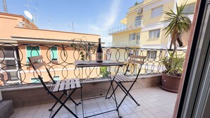 Un balcon avec une table et des chaises, un pot de fleurs et une vue sur les immeubles voisins.