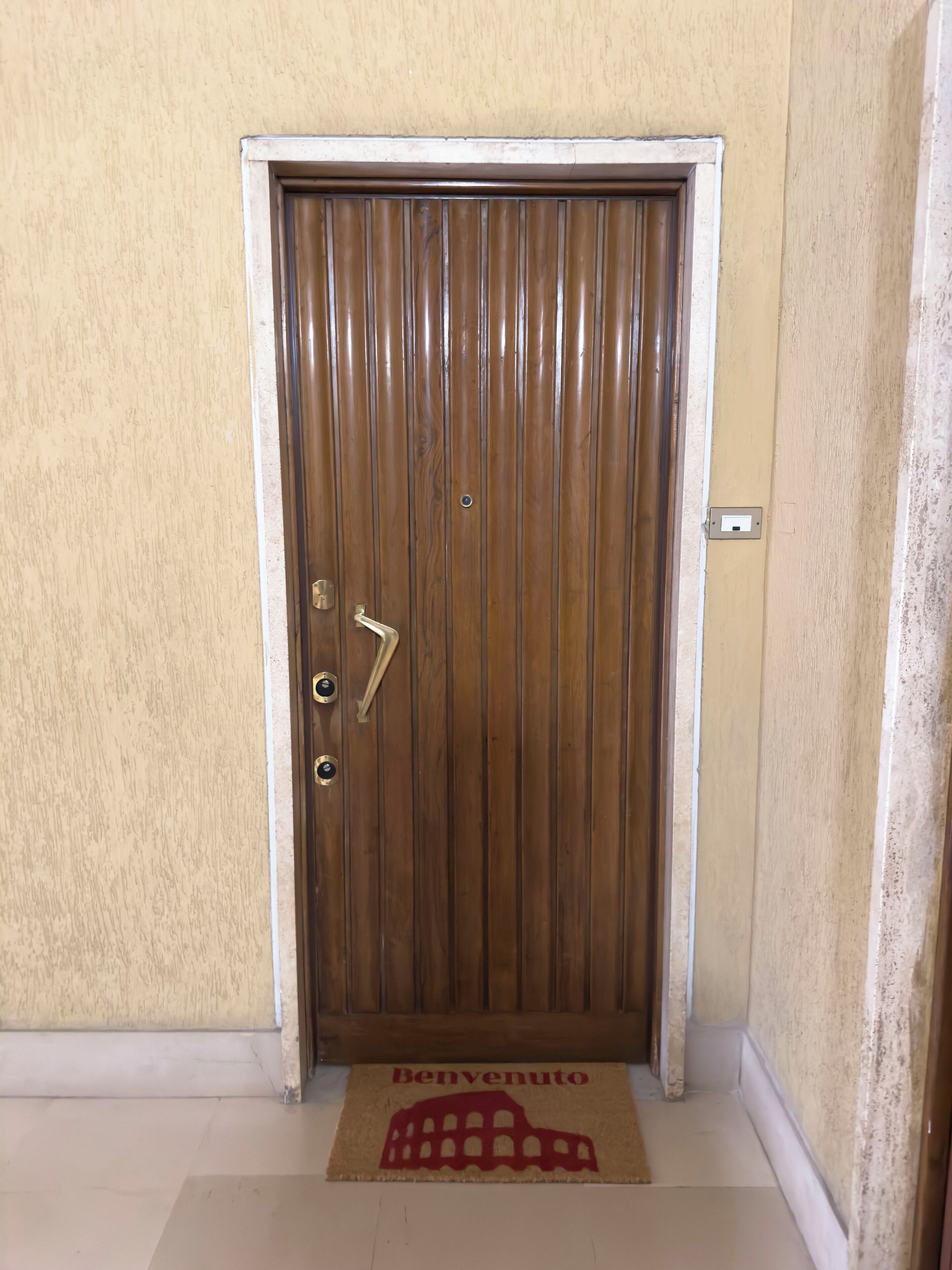 A wooden door with a brass handle and a keyhole, a doormat with "Benevento" and a coliseum design, and a beige wall with a white baseboard.