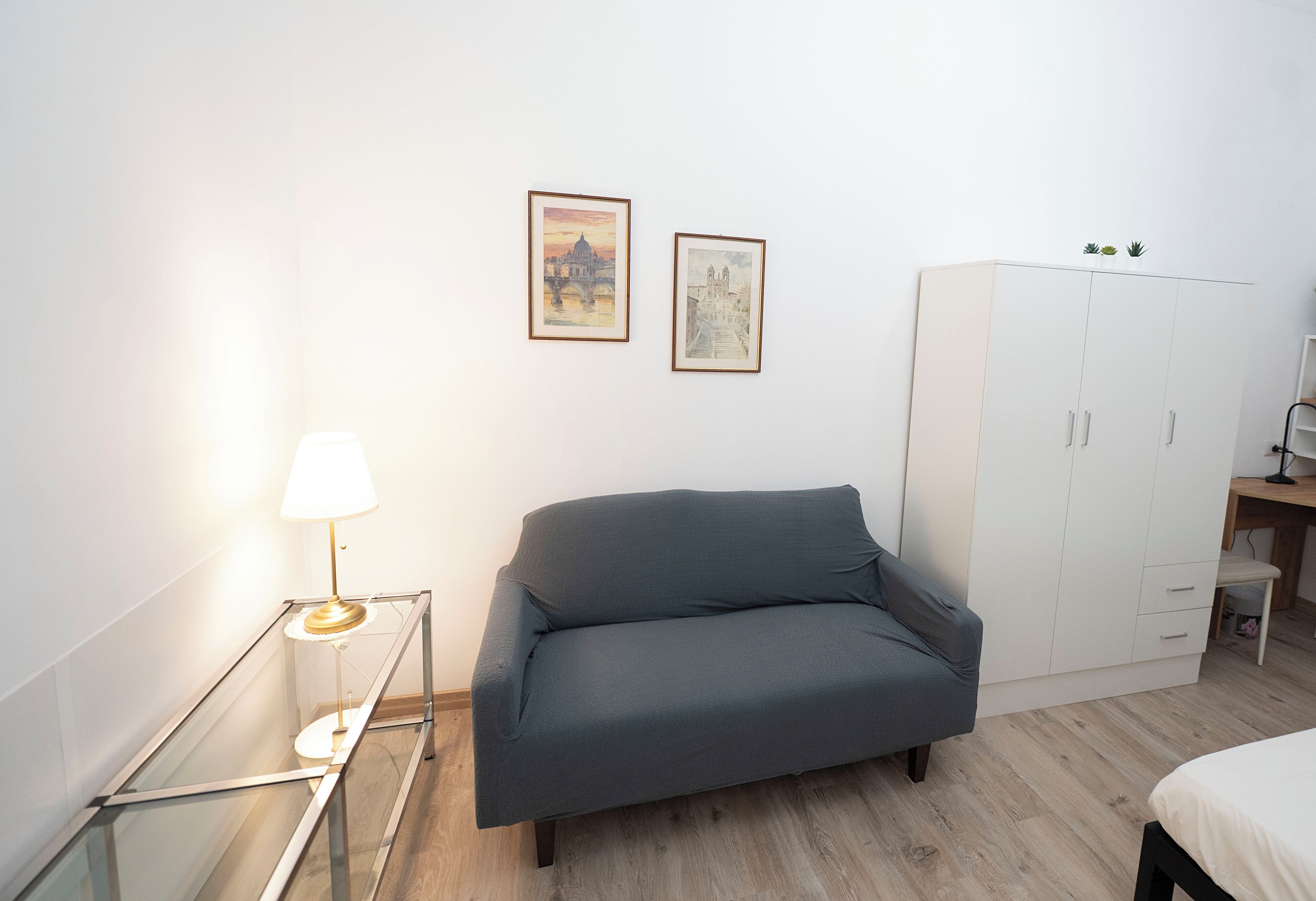 A room with a grey sofa, a glass table with a lamp, a white wardrobe, and two framed pictures on the wall.