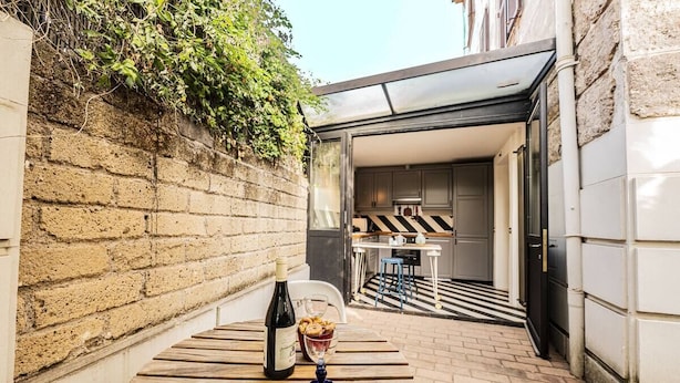 A narrow alleyway with a wooden table, a bottle of wine, and a glass. A kitchen is visible through an open door in the background.