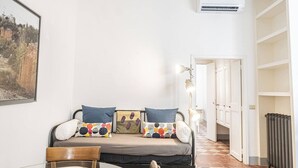 A cozy living room with a sofa, a dining table, and a chair. There is a framed picture on the wall, an air conditioning unit, and a radiator.