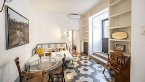 A compact living space with a dining area, a bed with colorful pillows, a wooden chair, a glass table, and a patterned floor.