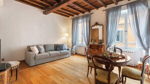 A living room with a wooden ceiling, a grey sofa, a dining table with chairs, and a window with curtains. - Monti  Apartments (Rome)
