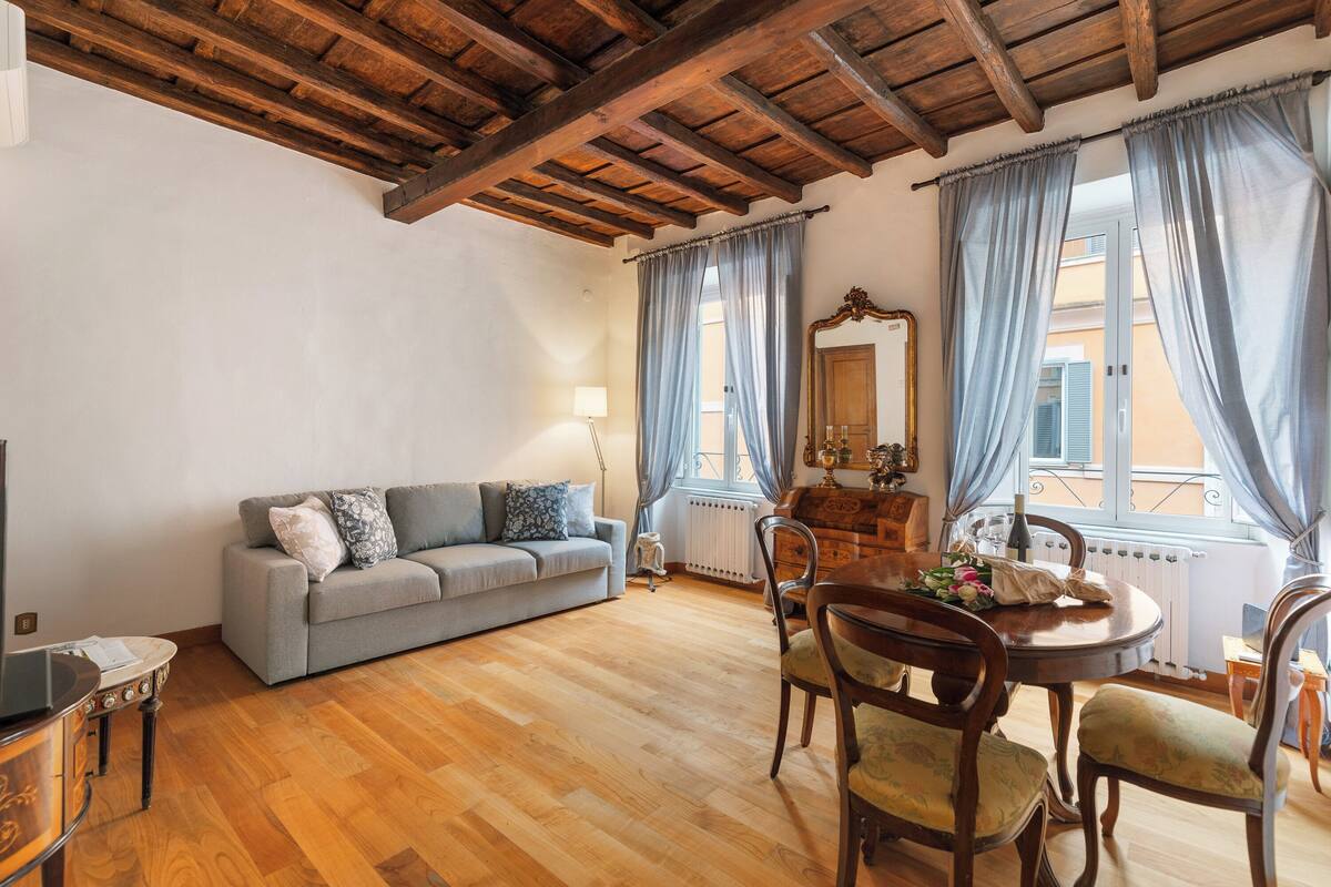 A living room with a wooden ceiling, a grey sofa, a dining table with chairs, and a window with curtains.