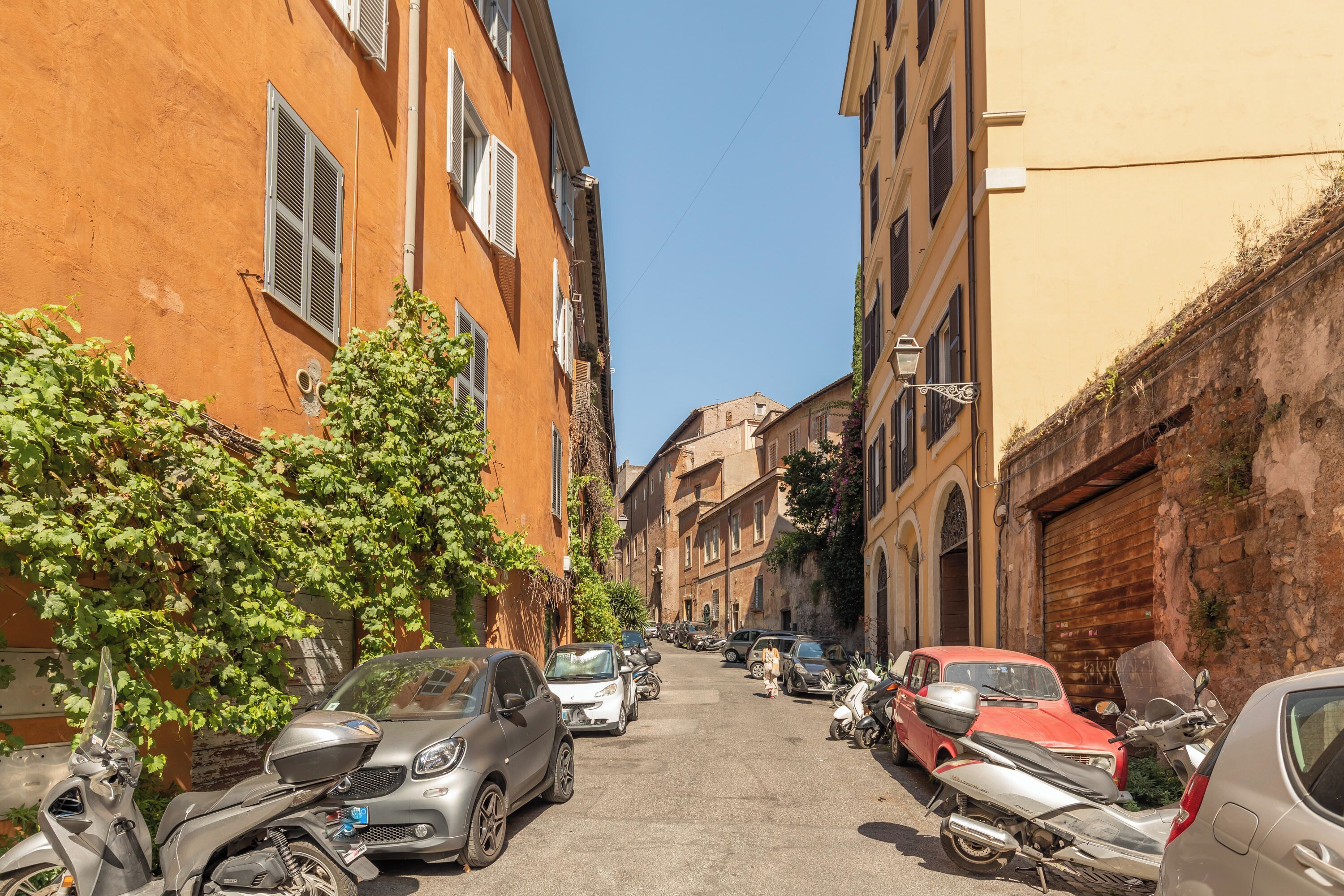 Una stretta via fiancheggiata da auto parcheggiate e scooter, edifici con persiane e un cielo azzurro intenso.