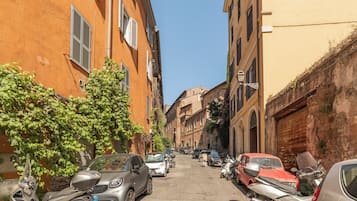 Una stretta via fiancheggiata da auto parcheggiate e scooter, edifici con persiane e un cielo azzurro intenso.