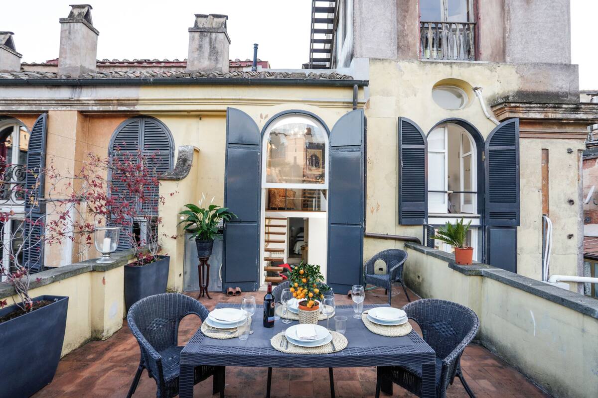 A rooftop terrace with a table set for dining, surrounded by potted plants and wicker chairs.