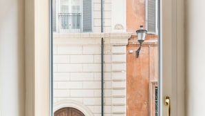 Una habitación con un ventanal grande, un banco con dos cojines estampados y vistas a un edificio con puerta y contraventanas.