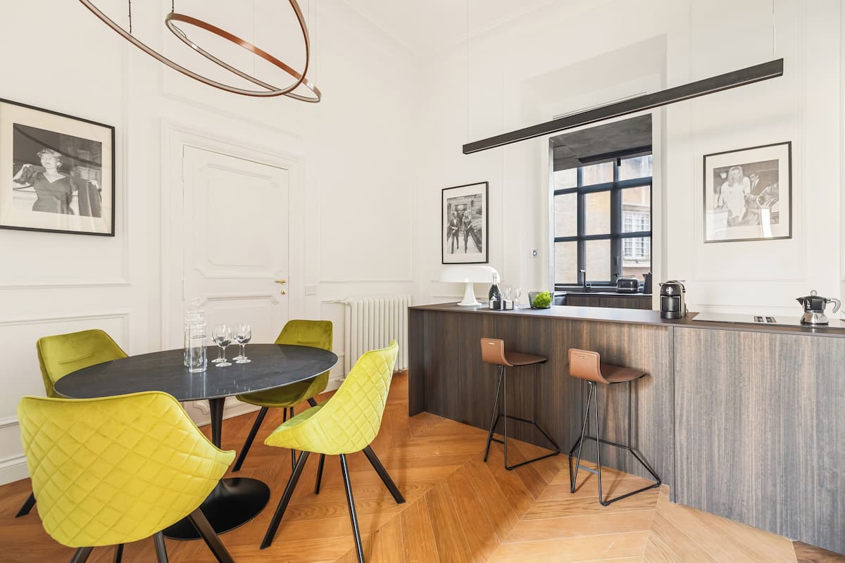 A modern kitchen with a central island, bar stools, and a dining area with a round table and yellow chairs.