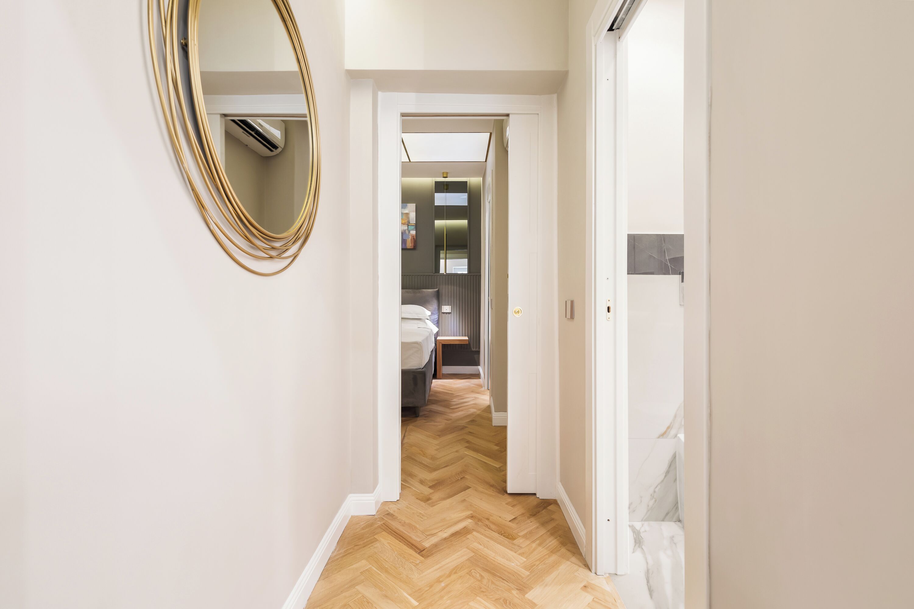 Un pasillo con suelo de espiga, un espejo y una puerta que da a una habitación con cama y mesita de noche.