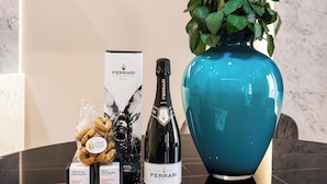 Una mesa con una botella de vino, una caja de bombones Ferrari, un florero con flores blancas y una revista.