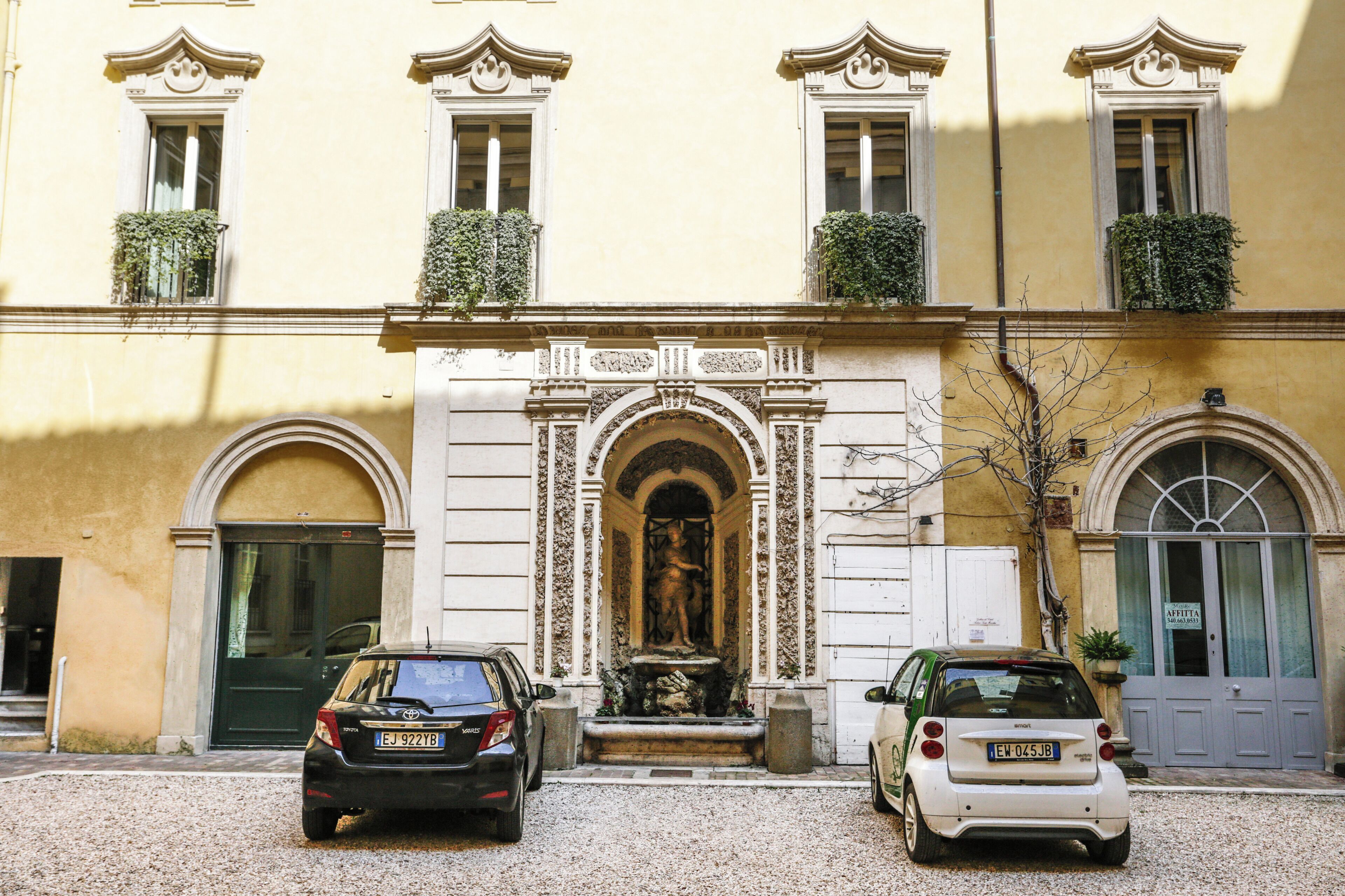 A building with a central arched entrance, two cars parked in front, and a statue in the doorway.