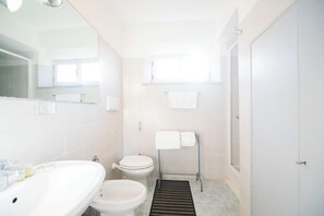 Une salle de bain équipée d’un lavabo, de toilettes et d’un bidet. Un miroir se trouve au-dessus du lavabo et une fenêtre au-dessus des toilettes.