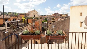 Una terraza con macetas de terracota con plantas, una barandilla de metal y vistas a un edificio histórico con una cruz en la parte superior.