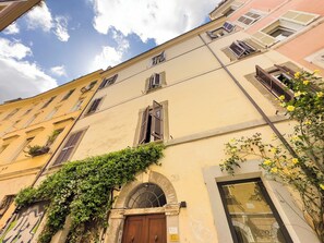 A building with multiple windows, some with shutters, and a door with an arch. There is greenery growing on the wall and a sign with text. - Trastevere Apartments (Rome)