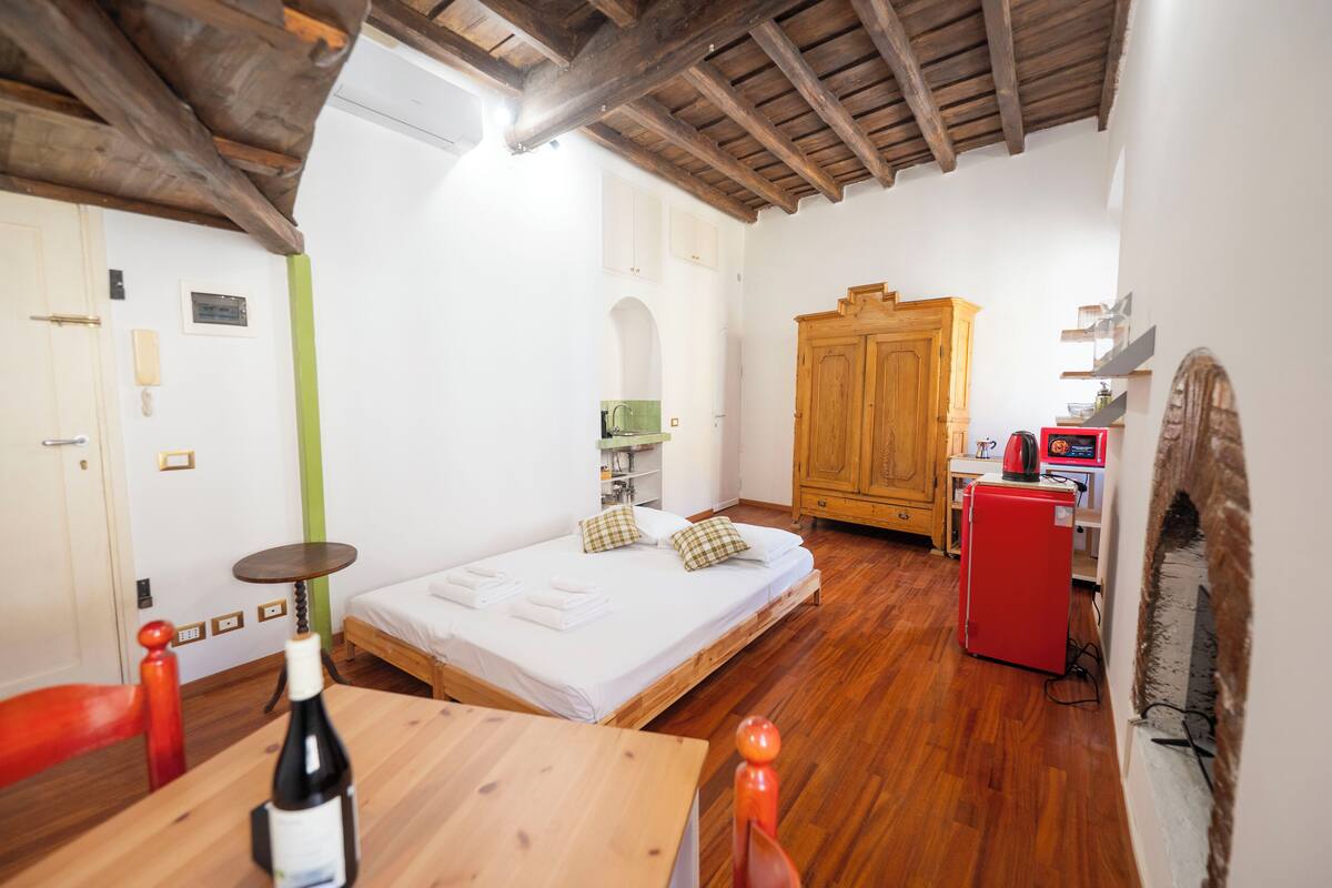 A bedroom with a wooden bed, a small table with a bottle, a red refrigerator, and a wooden cabinet.