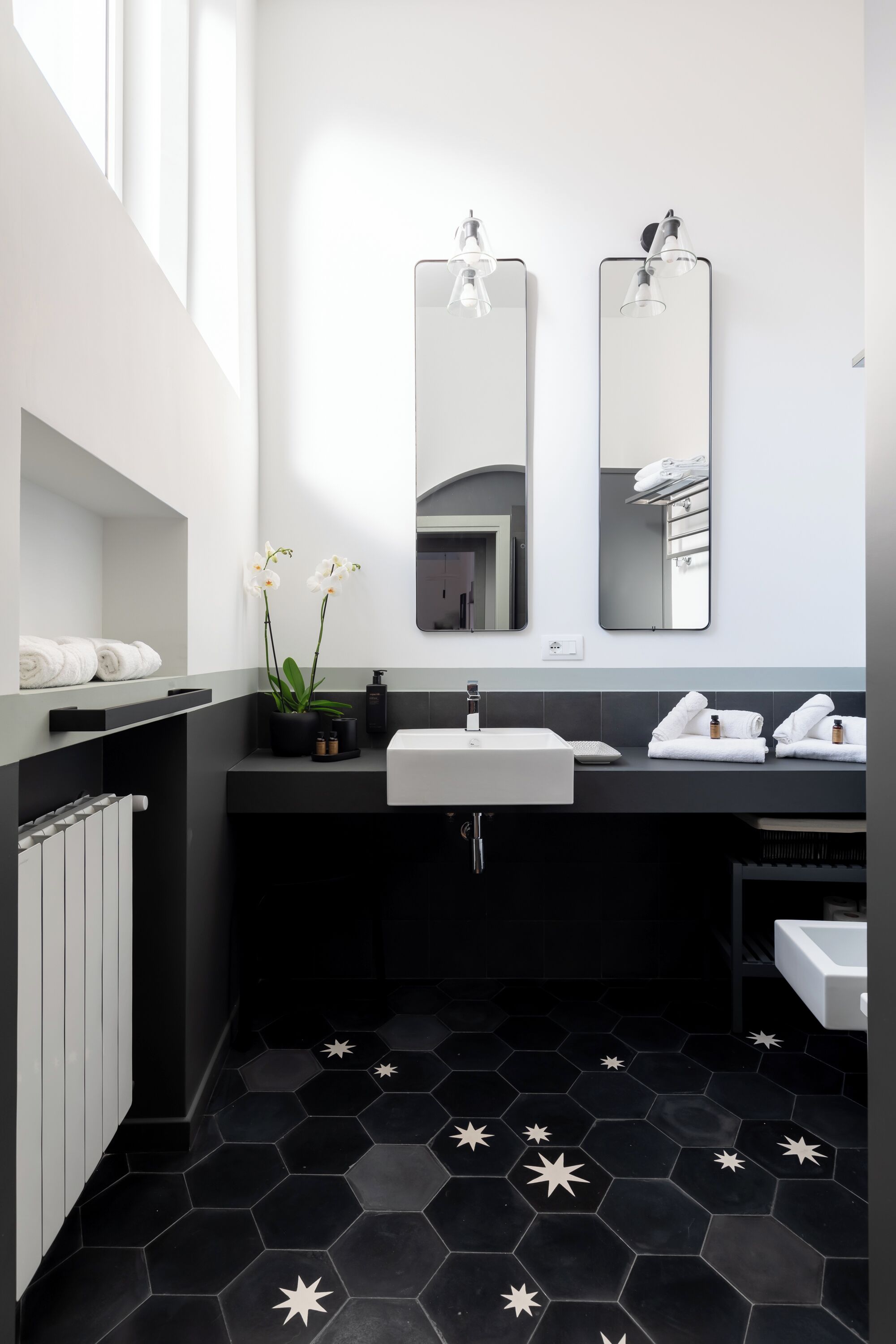 A modern bathroom with a black and white color scheme, featuring two sinks, a large mirror, and a black hexagonal tiled floor.