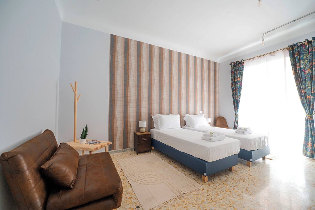 A hotel room with two beds, a brown leather sofa, a small wooden table with a lamp, and a striped wall.