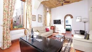 Una sala de estar con sofá, mesa de centro y televisor. Hay cortinas, ventilador de techo y ventana con vista al exterior.