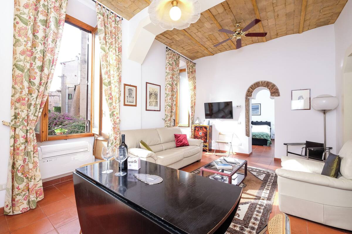 A living room with a sofa, coffee table, and a TV. There are curtains, a ceiling fan, and a window with a view of the outside.