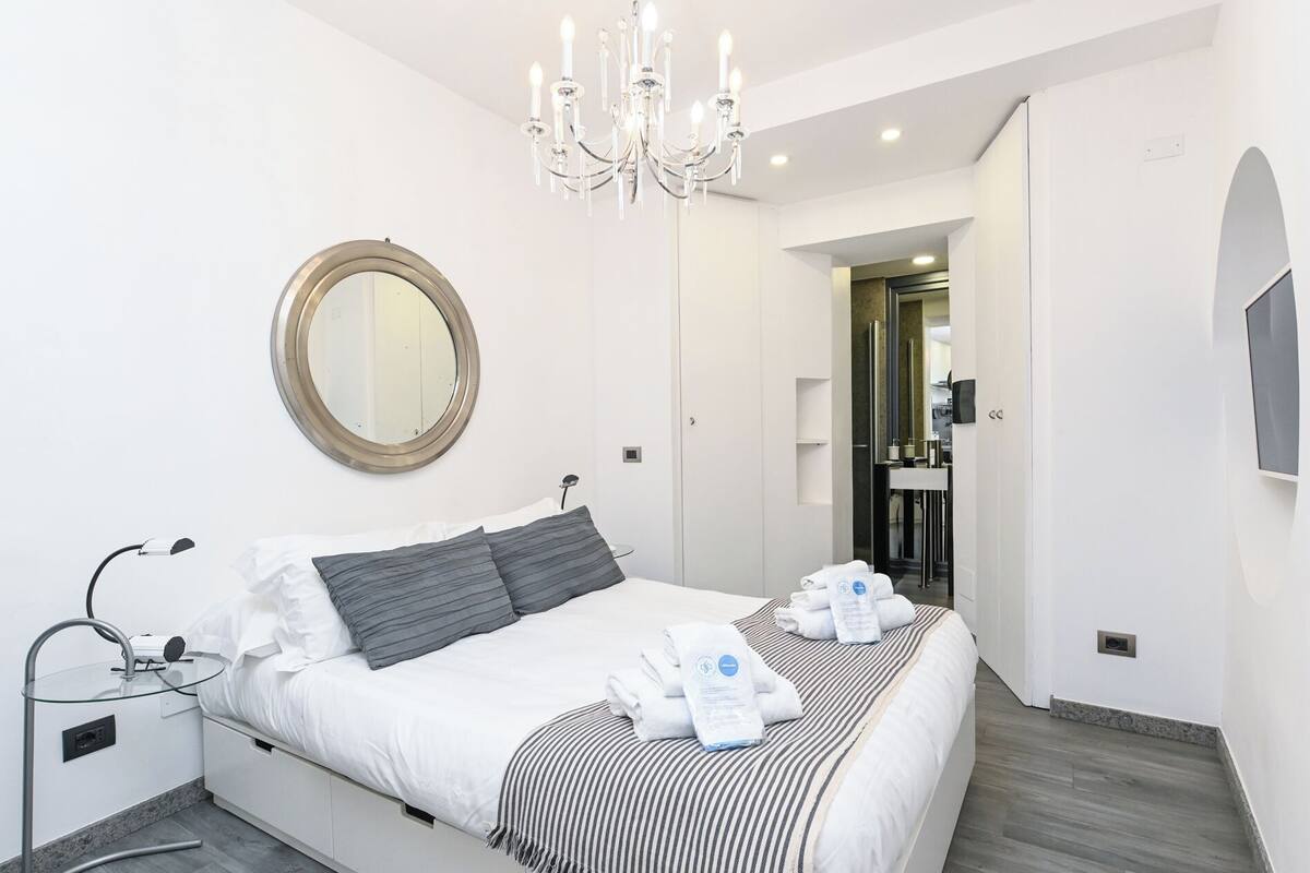 A modern hotel room with a large bed, a round mirror, a chandelier, and a view into the bathroom.