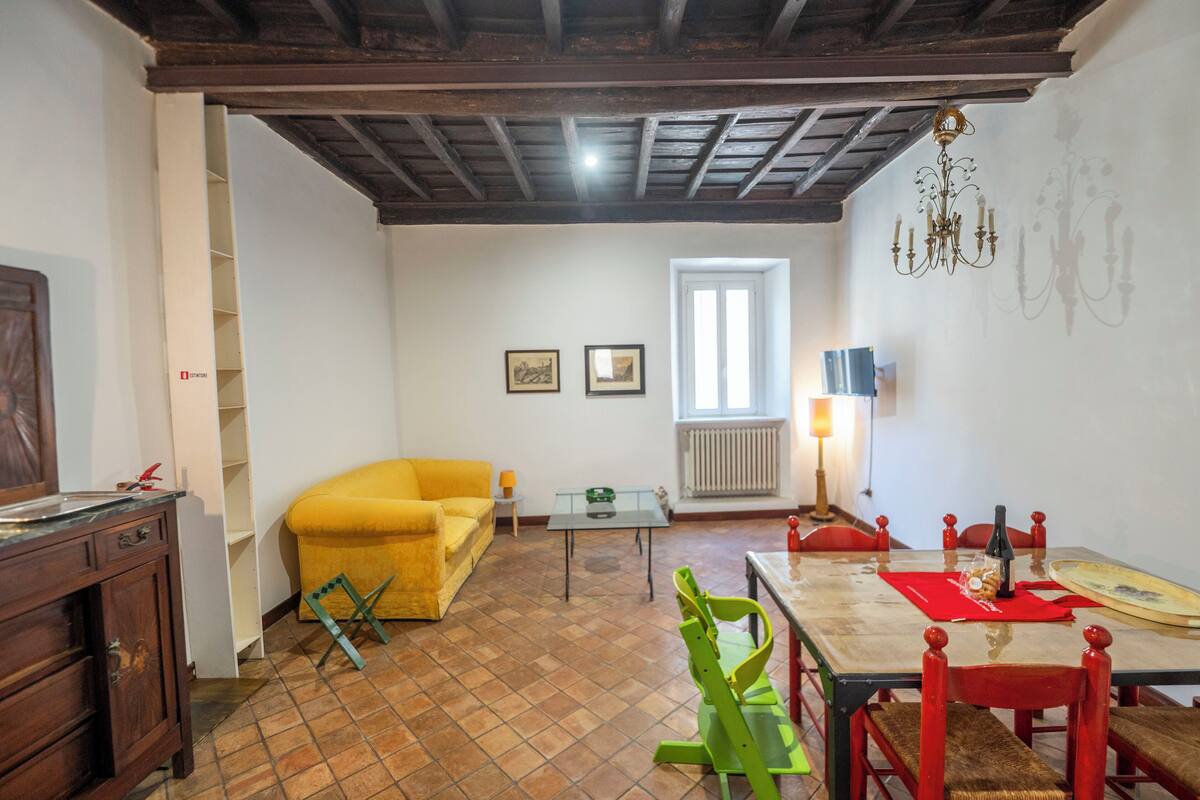 A dining area with a wooden table, red chairs, and a chandelier. There is a yellow sofa and a small table with a lamp in the background.