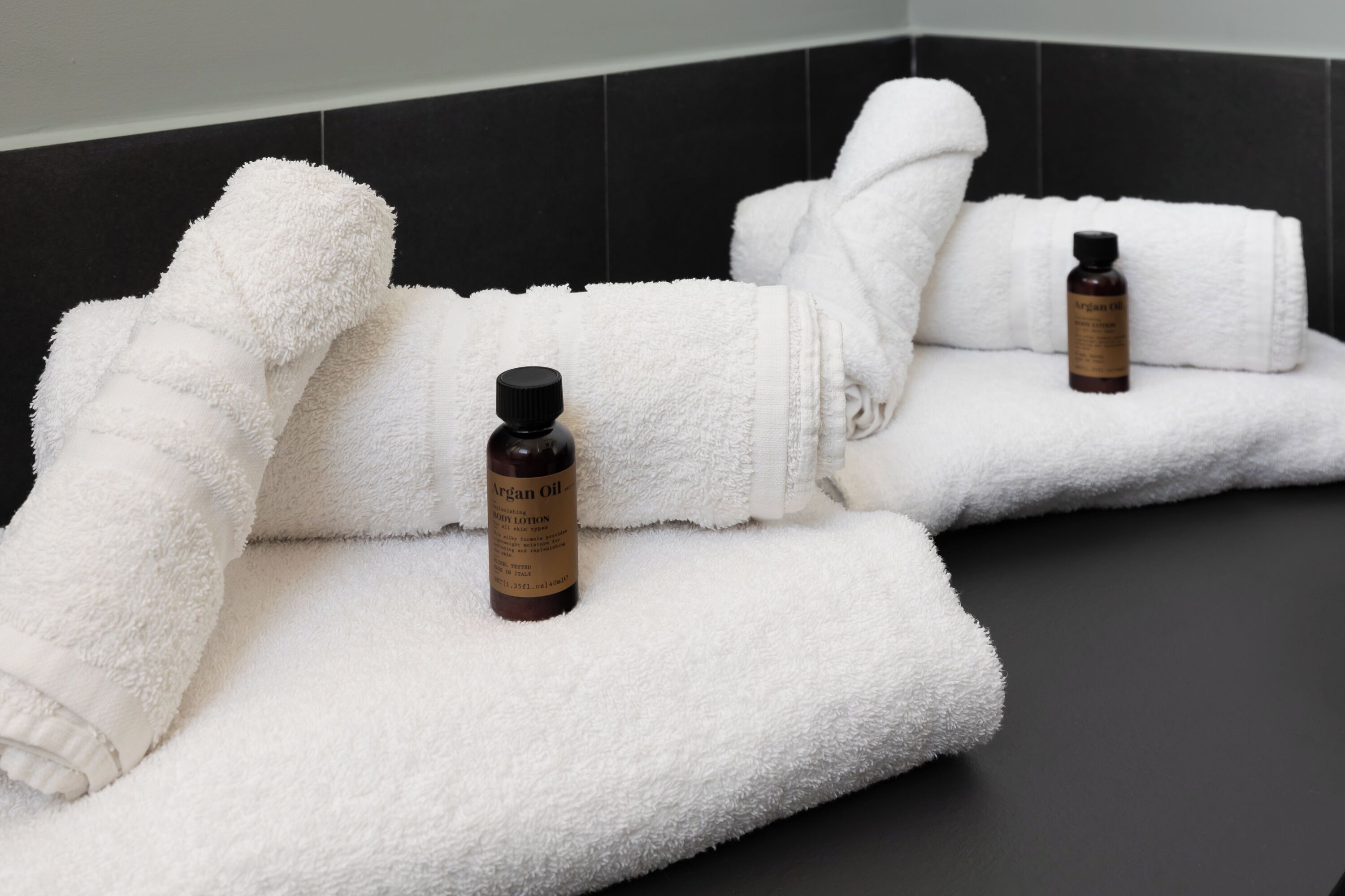 A bathroom with two white towels and two bottles of Argan Oil Body Lotion on a black tiled floor.