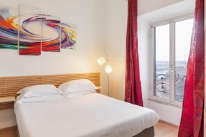 Ein Hotelzimmer mit einem Bett, einem großen Fenster mit roten Vorhängen und einem farbenfrohen, abstrakten Gemälde an der Wand.
