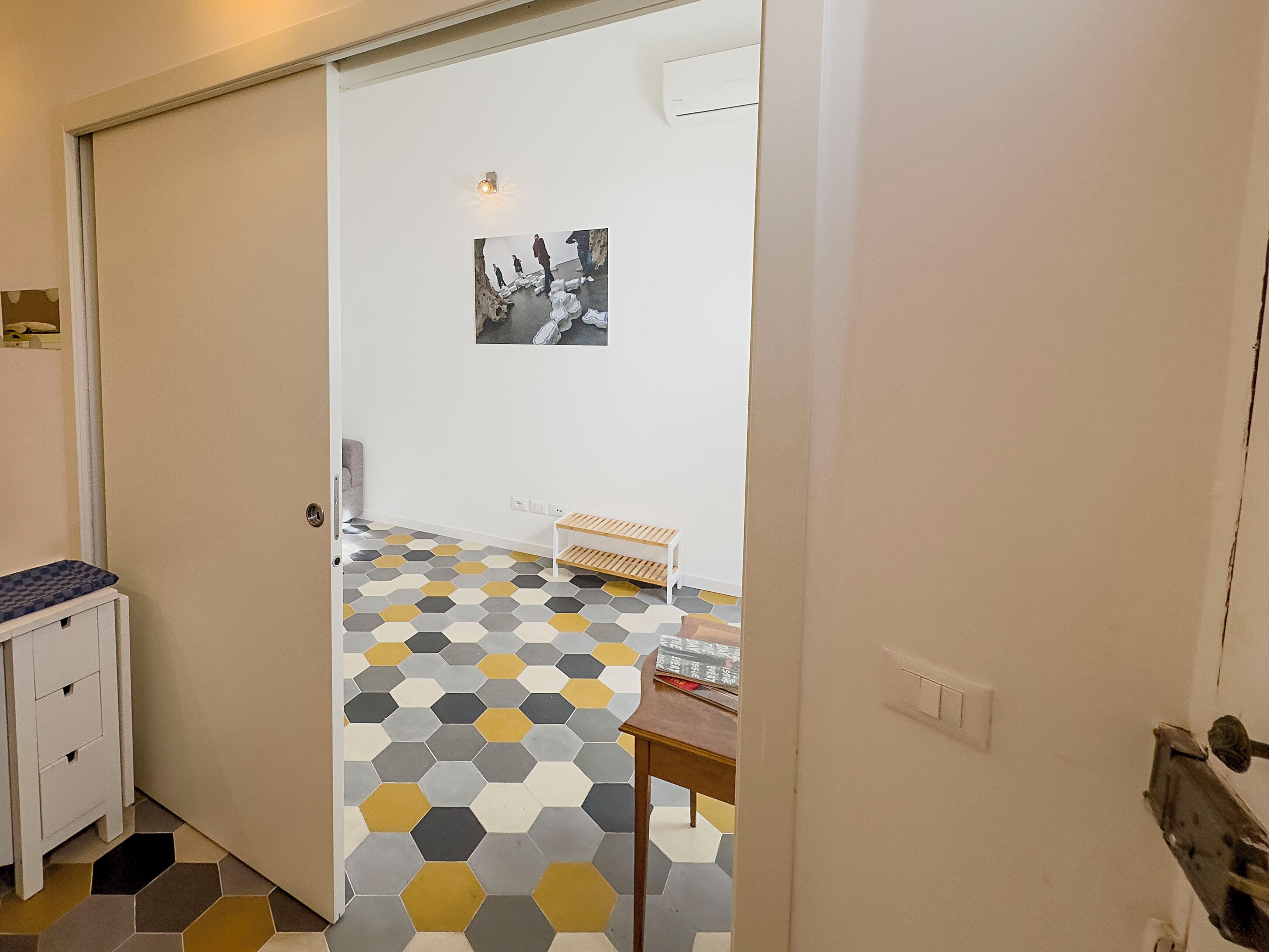 A hallway with a geometric patterned floor, a white door, a small table with a wooden top, and a wall-mounted air conditioning unit.
