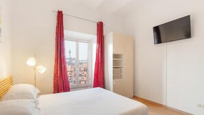 Ein Schlafzimmer mit einem Bett, einem an der Wand montierten Fernseher, einem Kleiderschrank und einem Fenster mit roten Vorhängen.