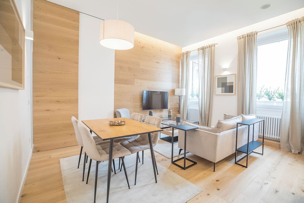 Un salón moderno con suelo de madera, una mesa de comedor con sillas, un sofá, un televisor y una ventana con cortinas.