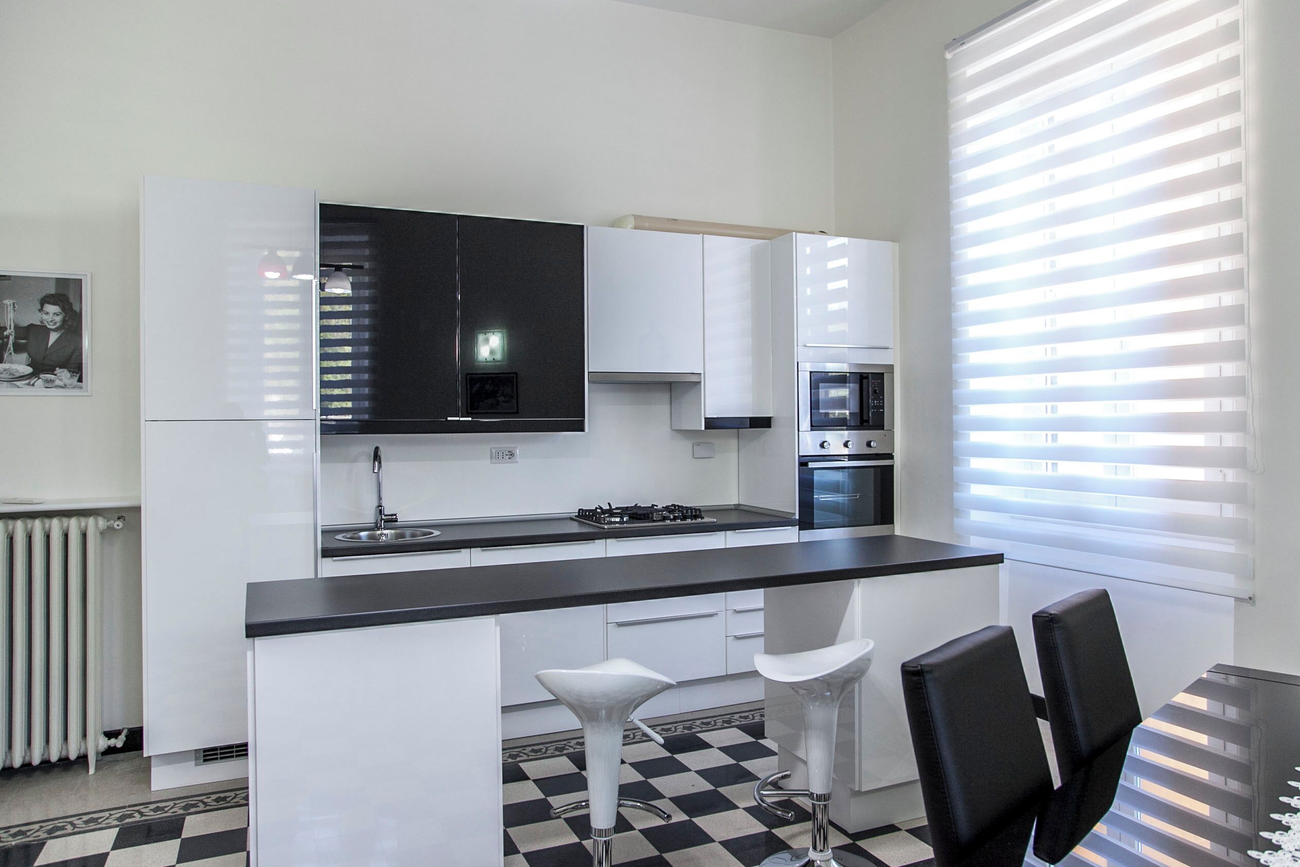 A modern kitchen with white cabinets, a black countertop, and a checkered floor. There are two bar stools and a dining table with chairs.